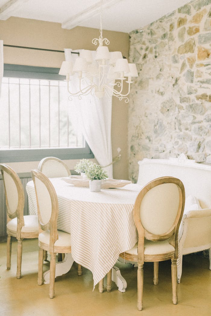 Charming dining room featuring a stone wall, white chandelier, and rustic wooden chairs.